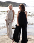 A woman stands on a sandy beach wearing the Arana Linen Pant in Natural. The ultra-wide leg flows gracefully in the breeze, complementing the relaxed, effortless silhouette. Soft sunlight highlights the lightweight gauze linen fabric, creating an airy and elegant look. The ocean waves gently roll in the background, enhancing the serene, coastal vibe.