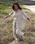 A woman stands on a sandy beach wearing the Arana Linen Pant in Natural. The ultra-wide leg flows gracefully in the breeze, complementing the relaxed, effortless silhouette. Soft sunlight highlights the lightweight gauze linen fabric, creating an airy and elegant look. The ocean waves gently roll in the background, enhancing the serene, coastal vibe.