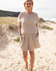 A woman stands barefoot on a sunlit beach, wearing the Olivia Linen Top in Off White, paired with Arana Linen Pants. The lightweight gauze linen fabric catches the ocean breeze, highlighting its soft, flowing texture. With gentle waves rolling in the background, the relaxed silhouette and natural tones create an effortlessly elegant coastal look.