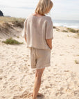 A woman stands barefoot on a sunlit beach, wearing the Olivia Linen Top in Off White, paired with Arana Linen Pants. The lightweight gauze linen fabric catches the ocean breeze, highlighting its soft, flowing texture. With gentle waves rolling in the background, the relaxed silhouette and natural tones create an effortlessly elegant coastal look.