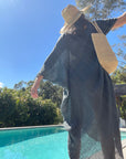 Person wearing a blue linen dress and raffia hat by a pool with trees in the background
