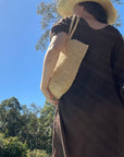 Woman wearing linen dress Raffia bag and hat outdoors with trees and blue sky in the background