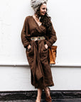 Woman in a brown outfit with a headscarf and sandals standing against a white wall.