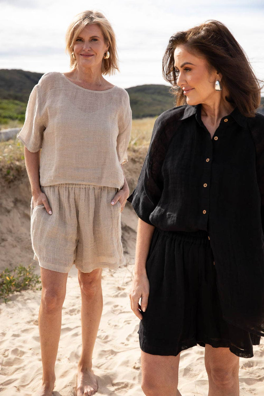 A woman stands barefoot on a sunlit beach, wearing the Olivia Linen Top in Off White, paired with Arana Linen Pants. The lightweight gauze linen fabric catches the ocean breeze, highlighting its soft, flowing texture. With gentle waves rolling in the background, the relaxed silhouette and natural tones create an effortlessly elegant coastal look.