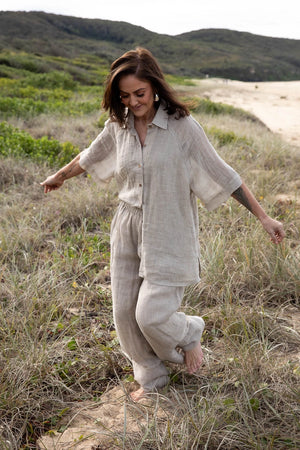 A woman stands on a sandy beach wearing the Arana Linen Pant in Natural. The ultra-wide leg flows gracefully in the breeze, complementing the relaxed, effortless silhouette. Soft sunlight highlights the lightweight gauze linen fabric, creating an airy and elegant look. The ocean waves gently roll in the background, enhancing the serene, coastal vibe.