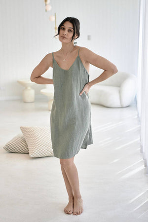 A woman poses in a minimalist studio setting, wearing the Linen Slip Dress in Pistachio. The soft, breathable 100% European linen drapes beautifully, highlighting the relaxed fit, scoop neckline, and adjustable straps. The subtle green hue enhances the dress’s fresh, modern aesthetic, perfect for effortless styling.