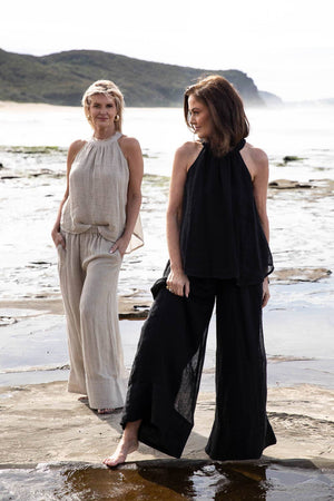 A woman stands on a sandy beach wearing the Arana Linen Pant in Natural. The ultra-wide leg flows gracefully in the breeze, complementing the relaxed, effortless silhouette. Soft sunlight highlights the lightweight gauze linen fabric, creating an airy and elegant look. The ocean waves gently roll in the background, enhancing the serene, coastal vibe.