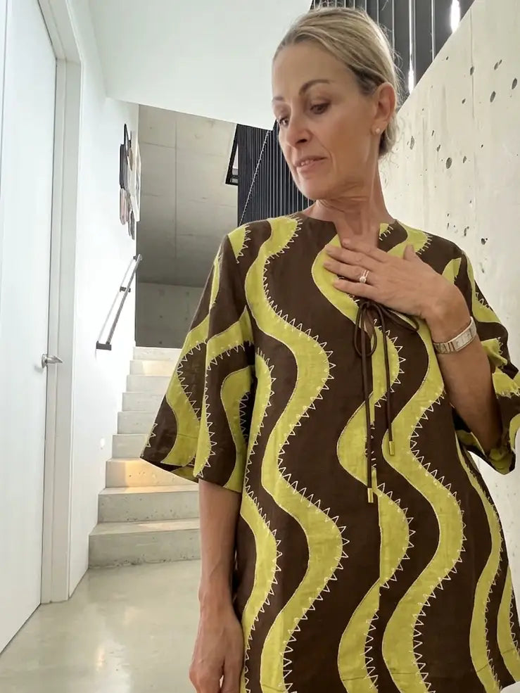 Alemais Linen Dress Worn by woman brown and yellow wavy patterned kaftan dress, standing in a modern interior hallway with concrete walls and stairs.