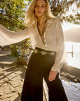 Woman in a whiteESTILO Emporio linen blouse and black pants standing by a body of water with trees in the background