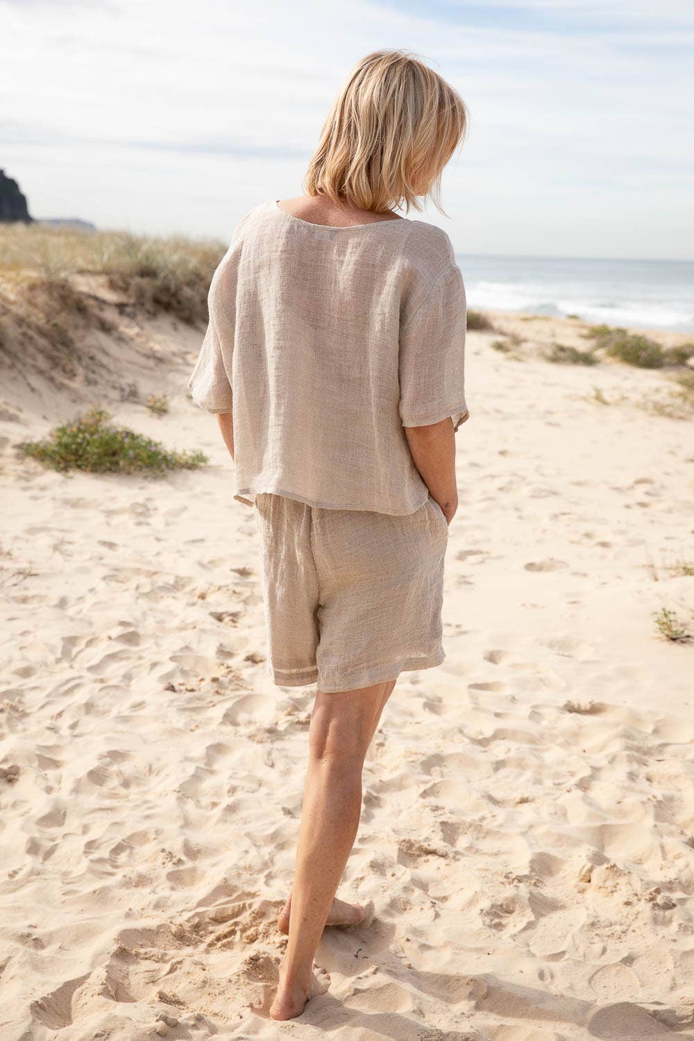 A woman stands barefoot on a sunlit beach, wearing the Olivia Linen Top in Off White, paired with Arana Linen Pants. The lightweight gauze linen fabric catches the ocean breeze, highlighting its soft, flowing texture. With gentle waves rolling in the background, the relaxed silhouette and natural tones create an effortlessly elegant coastal look.