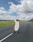 Woman in a white linen dress and hat standing on a road with a blue sky and clouds in the background