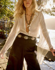 Woman wearing a white Estilo Emporio linen blouse and black pants with a belt, standing outdoors near water.