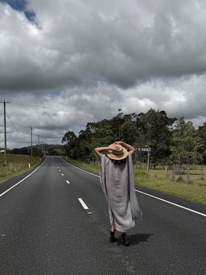Person in a Estilo Emporio dress and hat walking alone on a long, straight road under a cloudy sky.