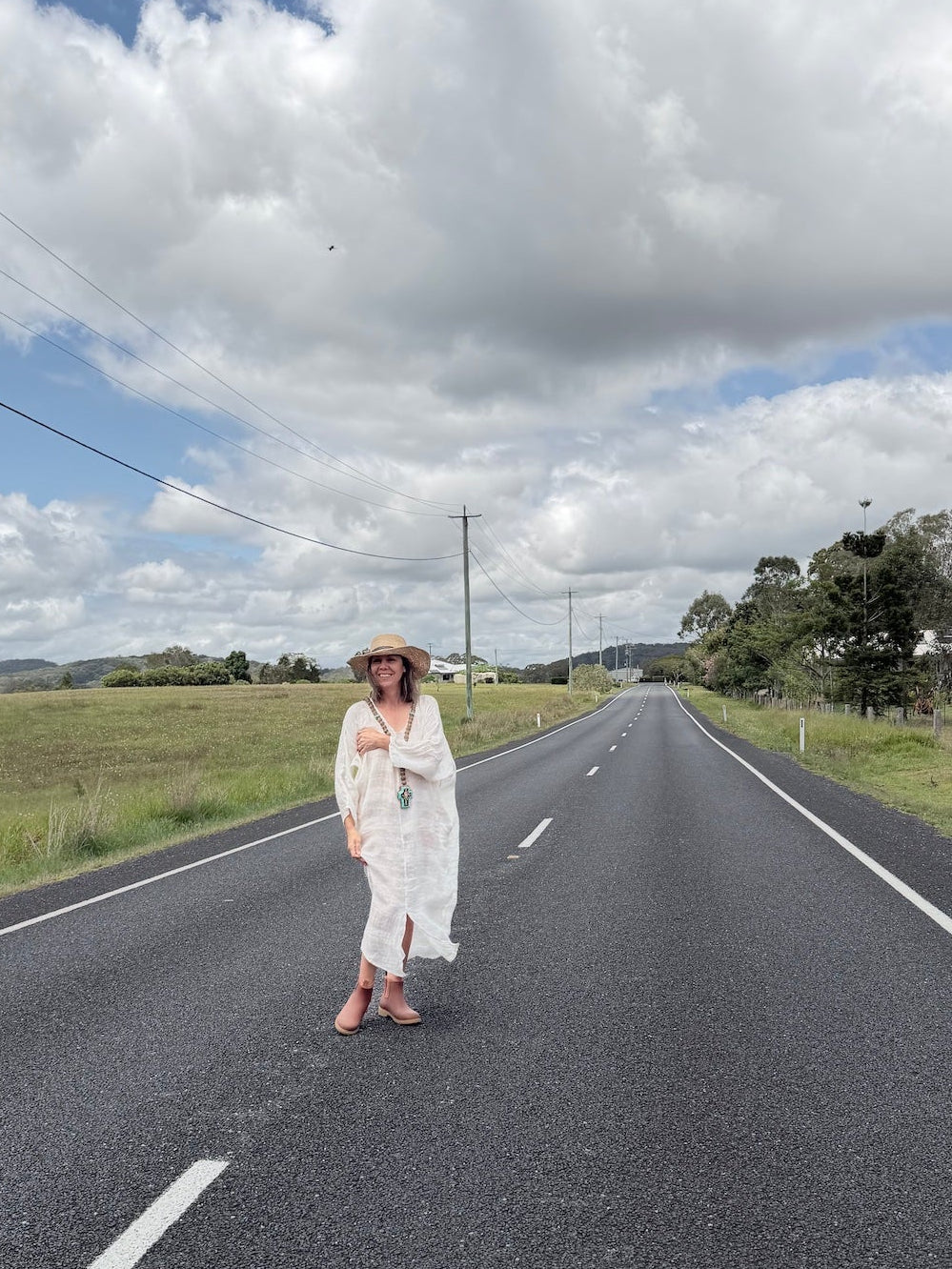 Person in a white linen dress standing on a rural road with a blue sky and clouds.