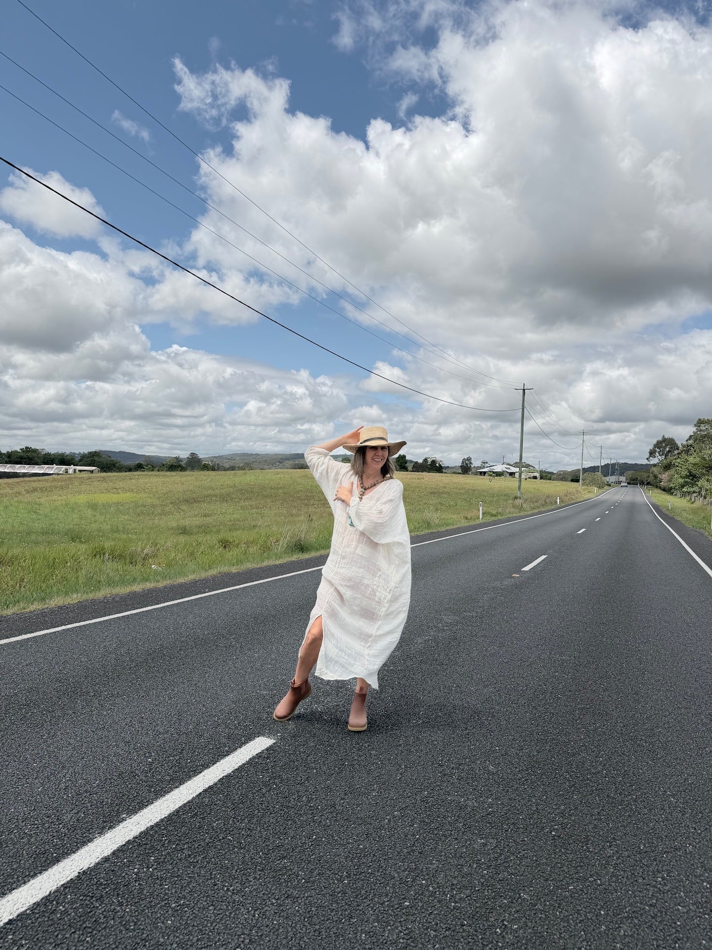 Woman in a white linen dress and hat standing on a road with a blue sky and clouds in the background