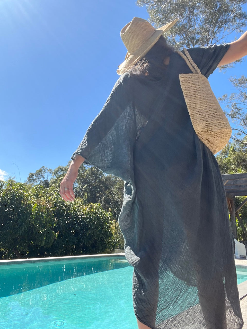 Person wearing a blue linen dress and raffia hat by a pool with trees in the background