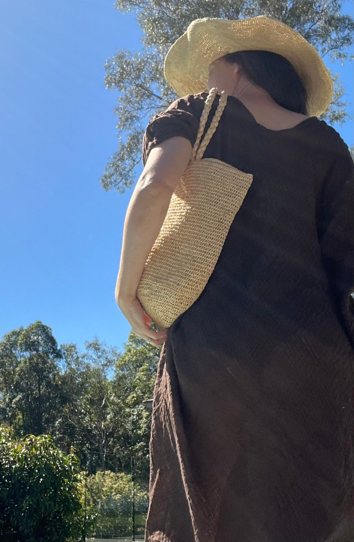 Woman wearing linen dress Raffia bag and hat outdoors with trees and blue sky in the background
