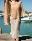 Person wearing a beige outfit standing outdoors on a concrete path with grass and trees in the background.