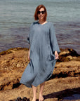 A woman La Bottega di Brunella Linen Dress, long-sleeve blue-gray textured  and sunglasses stands barefoot on rocky shoreline with water in the background. A pair of black sandals rests on the rocks beside her.