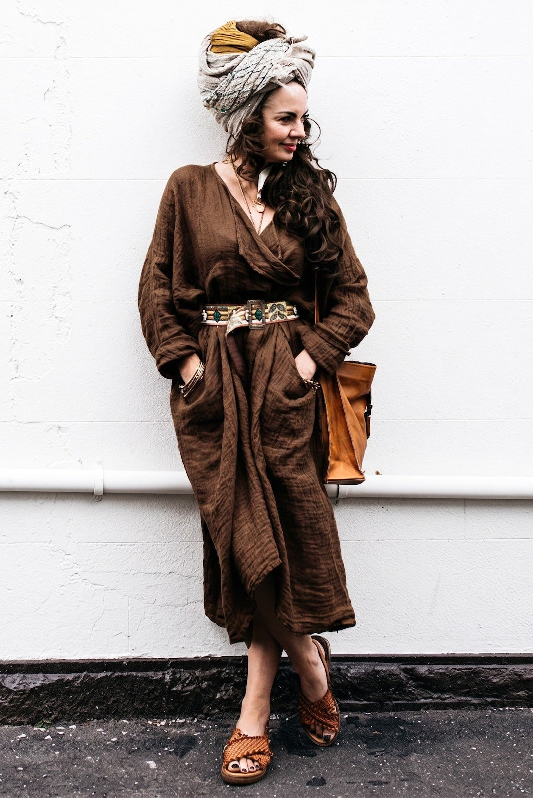 Woman in a brown outfit with a headscarf and sandals standing against a white wall.