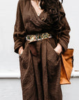 Person wearing a brown outfit with a belt and holding a brown bag against a light background