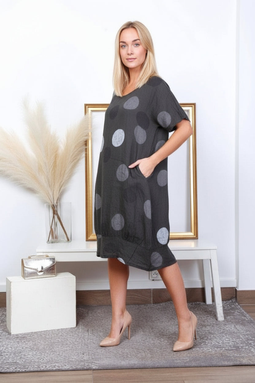 Woman wearing a black dress with gray polka dots in a room with a white wall and a small table.
