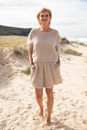 A woman stands barefoot on a sunlit beach, wearing the Olivia Linen Top in Off White, paired with Arana Linen Pants. The lightweight gauze linen fabric catches the ocean breeze, highlighting its soft, flowing texture. With gentle waves rolling in the background, the relaxed silhouette and natural tones create an effortlessly elegant coastal look.