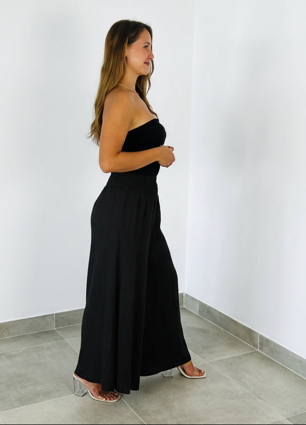 Woman wearing a black strapless jumpsuit against a white wall.